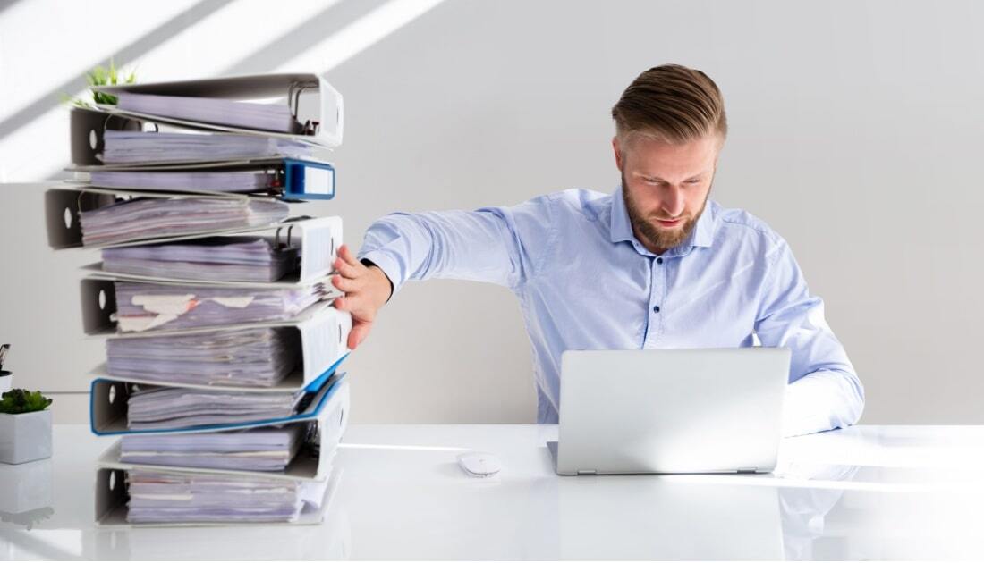 Business professional working on a laptop with a large stack of paper binders beside him, illustrating the benefits of electronic records in reducing paperwork and improving efficiency.