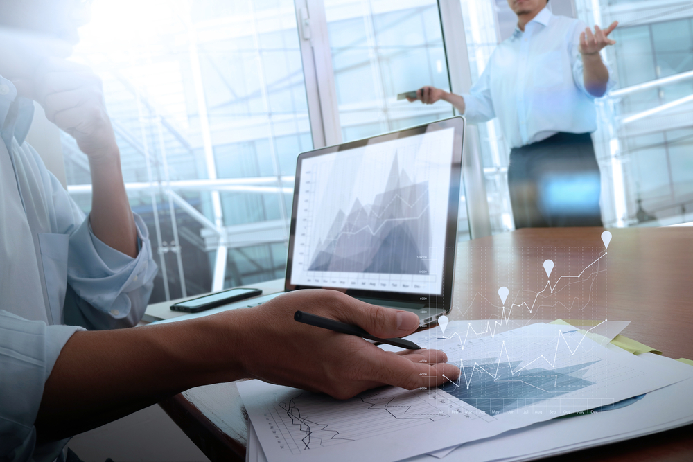 business documents on office table with smart phone and laptop computer and graph business with social network diagram and two colleagues discussing data in the background